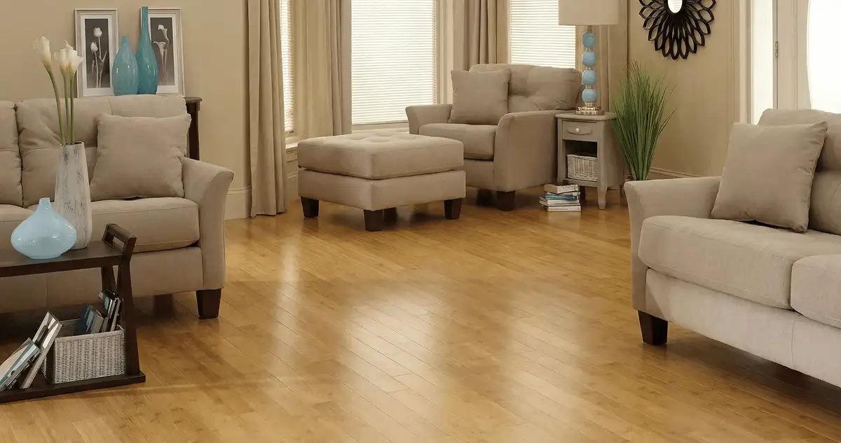 A living room with professionally installed bamboo flooring, beige sofas, a matching ottoman, a side table with a blue lamp, vases, framed pictures, and large windows with blinds and curtains in Bloomington, IN